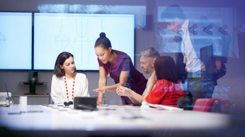 People gather around a table in conversation, with their focus on a screen on the table