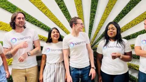 7 of the Mentra employees wearing branded Mentra t-shirts against a background with colorful yellow and green strips.