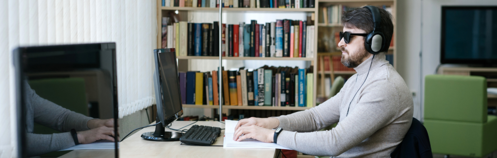 A blind man at their desk wearing a headset and reading a book in braille.