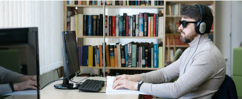 A blind man at their desk wearing a headset and reading a book in braille.