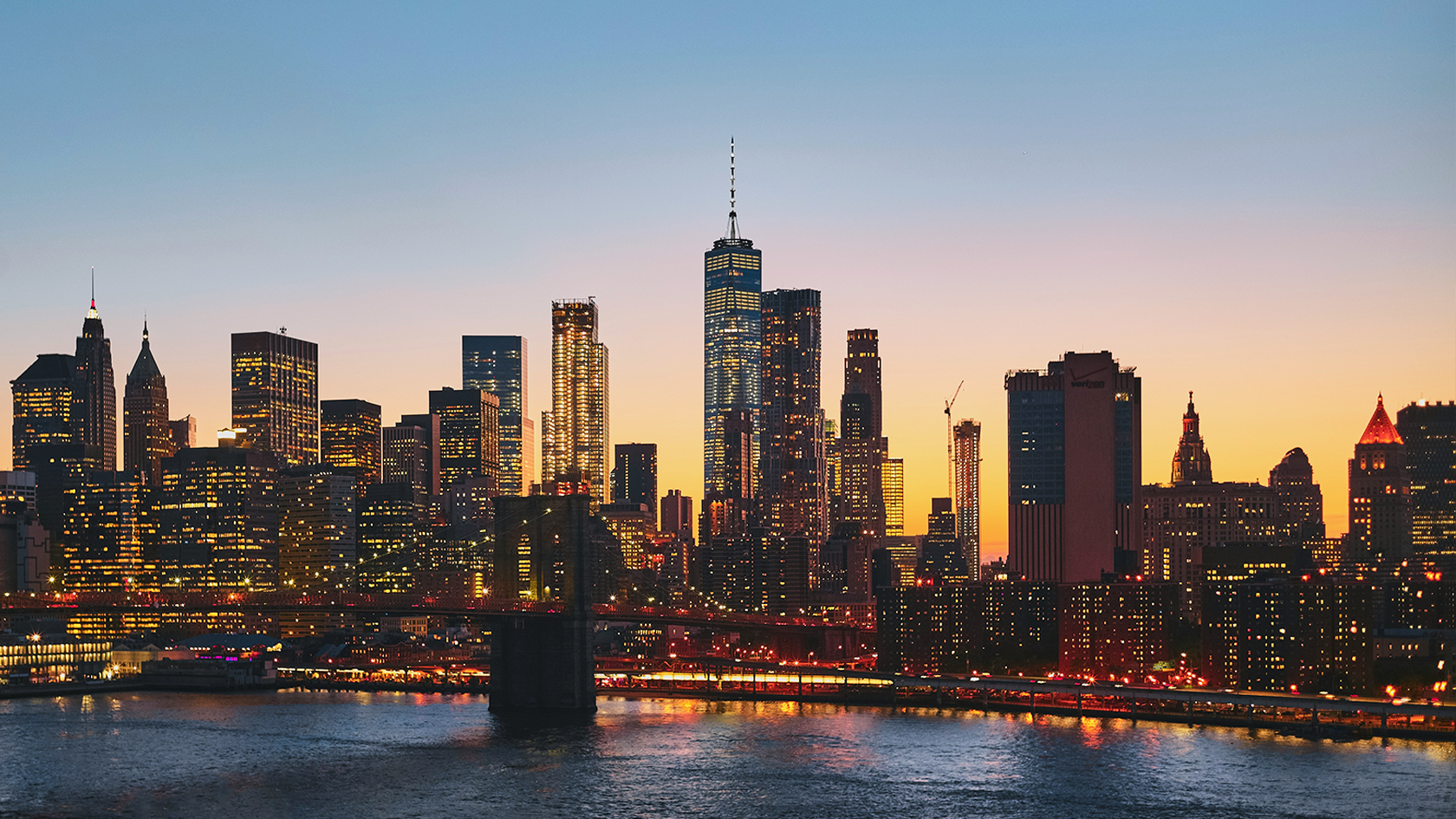 A vibrant city skyline at dusk, featuring a variety of illuminated skyscrapers with a river in the foreground and a bridge spanning across. The sky is a gradient of warm oranges transitioning into a cool blue.